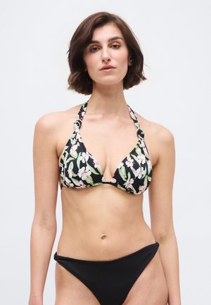 Νεαρή γυναίκα με κοντά καστανά μαλλιά, φορώντας μπλούζα μπικίνι με μαύρα λουλούδια και απλό κάτω μέρος μπικίνι σε μαύρο χρώμα, στέκεται μπροστά σε απλό φόντο.