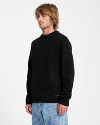 Man wearing a black long-sleeve sweater and light blue washed jeans standing against a plain white background.