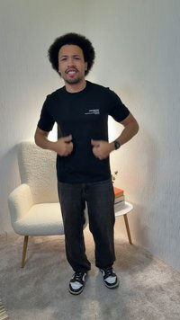 Mężczyzna z afro, ubrany w czarną koszulkę i tenisówki, stoi uśmiechnięty w pokoju z wykładziną, obok białego fotela i małego okrągłego stolika z książkami.