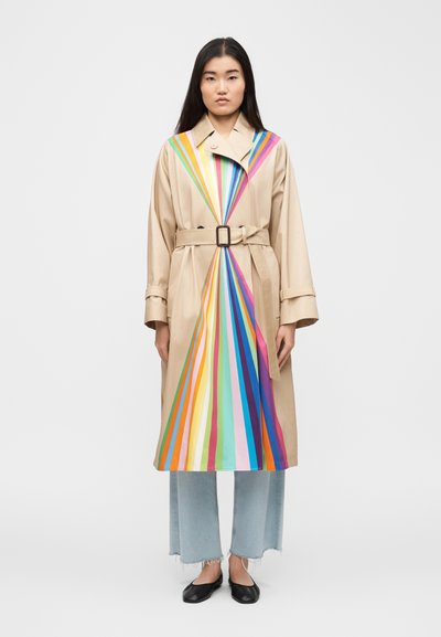 Woman stands wearing beige belted trench coat with colorful vertical stripes converging at waist, light blue cropped jeans, and black flats.