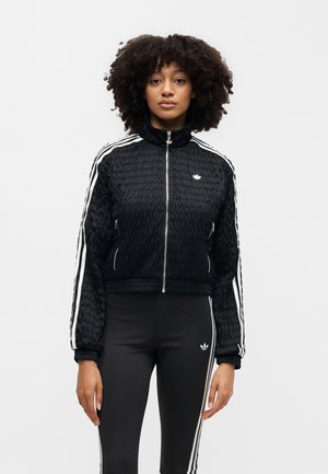 Žena s kučeravými vlasmi v čiernej bunde Adidas s bielymi pruhmi a čiernych legínach s rovnakými bielymi pruhmi stojaca pred bielym pozadím.