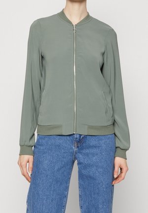 Personne portant une veste bomber vert olive clair avec fermeture éclair frontale et poignets côtelés, associée à un jean en denim bleu.