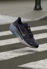 Dark blue Nike Air Zoom running shoe with white paint splatters hovering above a zebra crossing on asphalt.