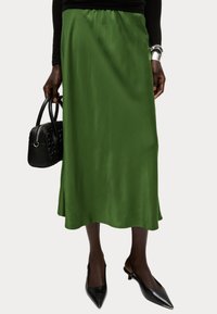 Grüne Satin-Midirock mit einem elastischen Bund, der eine glatte Textur und ein fließendes Design aufweist, kombiniert mit einer schwarzen Handtasche und High Heels.