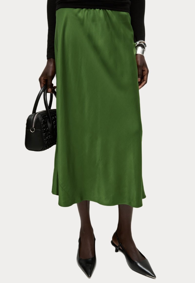 Grüne Satin-Midirock mit einem elastischen Bund, der eine glatte Textur und ein fließendes Design aufweist, kombiniert mit einer schwarzen Handtasche und High Heels.
