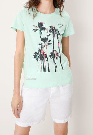 Žena s mátově zeleným tričkem s potiskem palmy a bílými šortkami stojí před jednobarevným světlým pozadím.