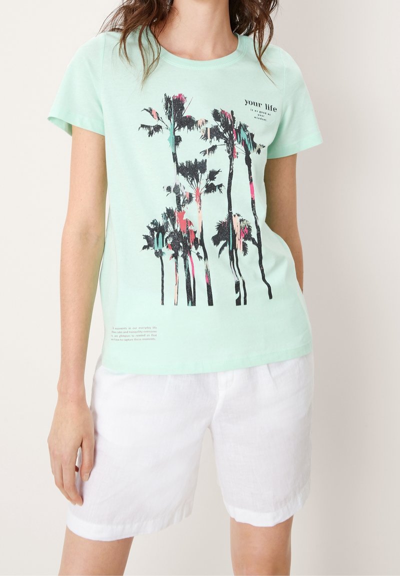 Femme portant un t-shirt vert menthe avec un motif de palmier et un short blanc, debout devant un fond clair uni.