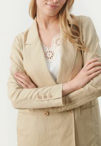 Beige blazer med skræddersyet design, der har blødt stof, dobbelte knapper foran og knapmanchetter. Bluse med blondeaccenter under.