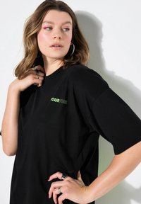 Černé oversized tričko vyrobené z bavlny, s nápisem "TVŮJ OHEŇ" v jasně zelené barvě. Krátké rukávy a uvolněný střih. Hladká textura.