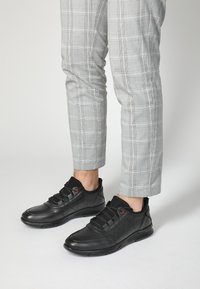 Schwarze Sportschuhe aus strukturiertem Leder, mit roten Ösen und einer gepolsterten Sohle, kombiniert mit grauen karierten Hosen.