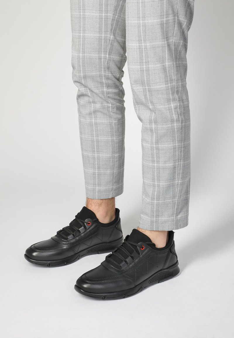 Schwarze Sportschuhe aus strukturiertem Leder, mit roten Ösen und einer gepolsterten Sohle, kombiniert mit grauen karierten Hosen.