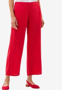 Femme portant un pantalon rouge ample avec un waistband élastique et des chaussures slingback à talon bas assorties, se tenant devant un fond blanc.