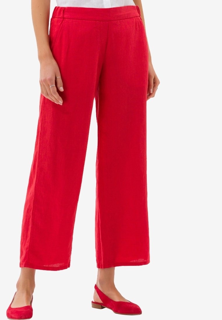 Vrouw in losse rode broek met een elastische tailleband en bijpassende rode lage slingback schoenen, staande tegen een witte achtergrond.