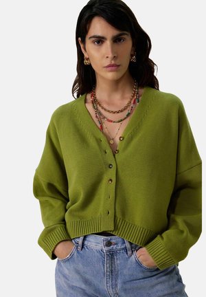 Green knitted cardigan with a V-neck, dropped shoulders, and button closure. Ribbed hem and cuffs. Worn over light blue jeans.