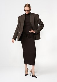 Barna szabott blézer egy testhezálló sötét ruhán; sima sziluettjét redőzött részletek és térd alatti hem jellemzik, fekete magassarkú cipőkkel párosítva.