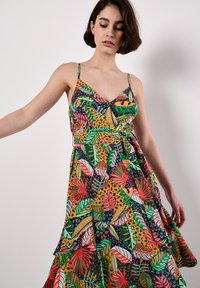 Model in einem bunten, gemusterten Kleid mit tropischen Blättern, das einen ausgestellten Saum hat. Sie wirkt gelassen und schaut mit einem subtilen Ausdruck nach unten.