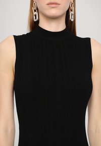 Rochie fără mâneci, neagră, cu un gât înalt. Cercei notabili din lanț cu detalii strălucitoare de aur și argint adaugă un contrast. Textură netedă.