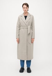 Donna in piedi, indossando un lungo cappotto beige con cintura, jeans blu e stivali neri spessi su uno sfondo bianco semplice.