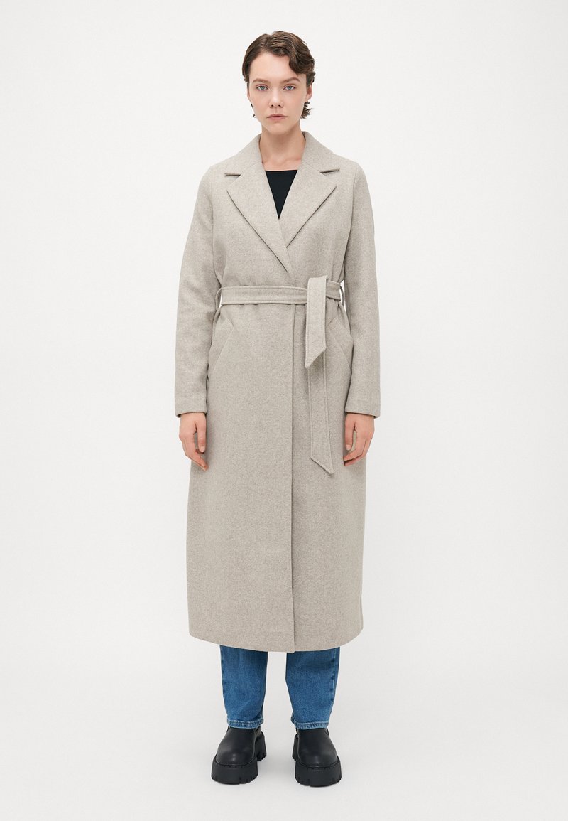 Donna in piedi, indossando un lungo cappotto beige con cintura, jeans blu e stivali neri spessi su uno sfondo bianco semplice.
