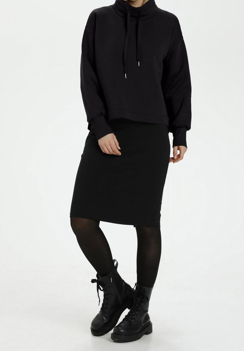 Černá krátká mikina s vysokým límcem a šňůrkami, v páru s černou přiléhavou sukní a černými kotníkovými botami na šněrování. Hladký povrch.