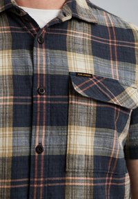 Chemise à manches courtes à carreaux en bleu marine, beige et orange. Présente un devant à boutons, une poche poitrine et une étiquette de marque discrète. En matériau coton.