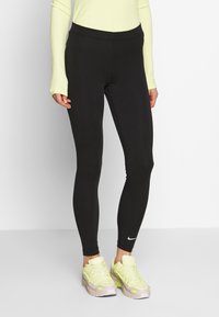 Svarta leggings med hög midja, tillverkade av stretchmaterial. Har en liten vit Nike-logotyp vid fållen. Kombineras med ljusa träningsskor.