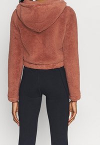 Sudadera corta de pelo sintético color óxido con una textura suave, que cuenta con capucha y hombros caídos, combinada con leggings negros.