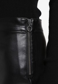 Närbild av svarta läderbyxor med en vertikal silverdragkedja och ringdragning, bärs med en svart ribbad tröja.