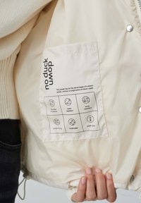Hellbeige Jacke aus einem glatten, wasserabweisenden Material. Enthält ein Etikett mit Symbolen für Isolierung, schnelles Trocknen und Atmungsaktivität.