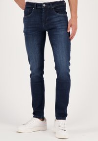 Dark blue, slim-fit denim jeans with five pockets, a zip fly, and a silver button. Paired with white sneakers. Smooth texture.