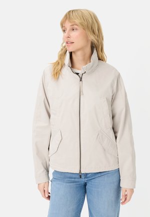 Kvinde iført lysebeige lynlåstrøje med høj krave og blå jeans, kigger til siden mod en hvid baggrund.