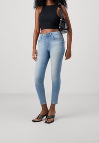 Slim-fit light blue jeans with a faded finish, paired with a black sleeveless crop top and black heeled flip-flops. Black handbag on shoulder.
