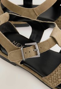 Sandale din suede bej cu o brățară pentru degete împletită și o talpă neagră netedă. Include o cataramă argintie ajustabilă pe brățara gleznei pentru o ajustare corespunzătoare.