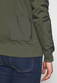 Olivově zelená mikina s texturovaným tělem z látky a lesklým, hladkým nylonovým rukávem a kapsou. Má detail na zipované boční kapse.