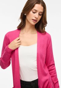 Fuchsia gebreide cardigan met lange mouwen en een open voorkant, gedragen over een witte top. Soepele textuur met geribde mouwen en details.