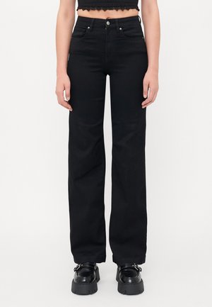 Person trägt schwarze, gerade geschnittene Hosen mit hoher Taille, vorderen Knöpfen und Reißverschluss, kombiniert mit schwarzen klobigen Plateauschuhen und einem kurzen schwarzen Oberteil.