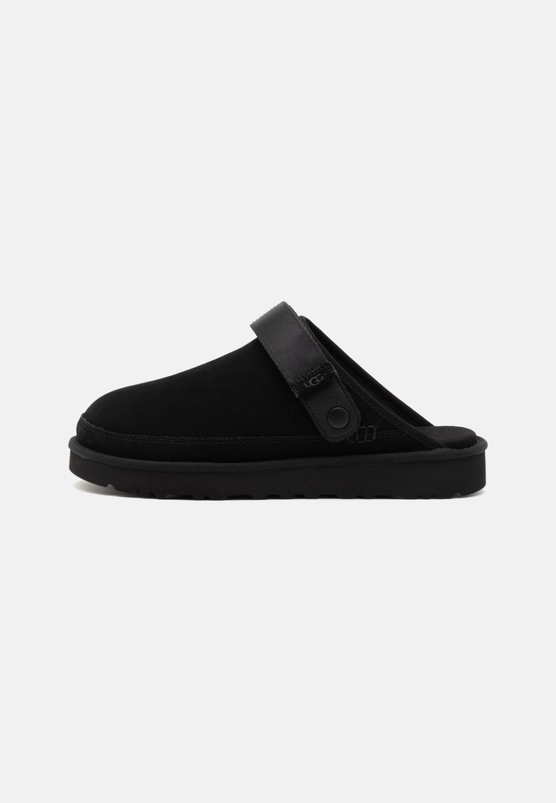 Black suede slip-on shoe with a wide strap featuring a circular flap. Thick black rubber sole and smooth texture. Minimalist design.