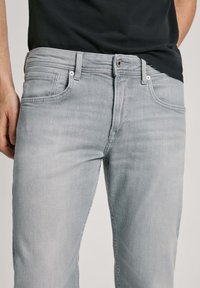 Hombre que lleva jeans de mezclilla gris claro y una camisa negra, mostrado desde la cintura hasta la mitad del muslo, de pie con los brazos relajados.