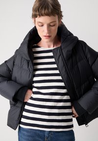 Doudoune noire avec col montant, associée à une chemise à rayures à manches longues en blanc cassé et bleu marine. Jean bleu visible à la taille.