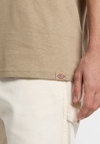 Beige t-shirt made of soft fabric, with short sleeves and a hem tag featuring the Dickies logo, worn with white cargo pants.