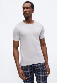 Άνδρας που φοράει ανοιχτό γκρι κοντομάνικη μπλούζα και μπλε-μαύρο καρό παντελόνι, κοιτάζοντας προς τα πλάγια σε λευκό απλό φόντο.