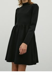 Abito nero a maniche lunghe con collo alto, corpetto aderente e gonna svasata, realizzato in un tessuto liscio con un aspetto leggermente strutturato.