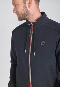 Marineblaue Zip-Überziehjacke mit hohem Kragen, silbernem Reißverschluss, roten Akzenten und Logo-Patch. Weicher Stoff und seitliche Taschen.