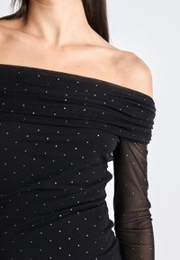 Femme portant une robe noire épaules dénudées avec des manches longues transparentes et de petits pois blancs sur le tissu.