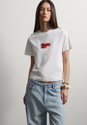 Junge Frau mit langen dunklen Haaren, die ein weißes T-Shirt mit einem roten Hummer-Design, hellblaue Jeans und ein Haargummi am Handgelenk trägt.