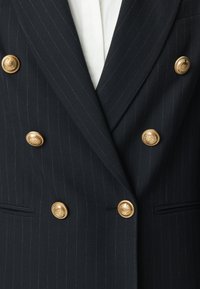 Marineblauw dubbelreihalig colbert met krijtstreep en zes gouden sierknopen, gedragen over een wit overhemd.