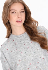 Pulover tricotat de culoare gri deschis, cu guler rotund și decorațiuni colorate răspândite, inclusiv mărgele mici în diferite culori.