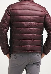 Vinröd vadderad jacka med horisontell quiltad design. Har svarta sidor och elastiska ärmmuddar, tillverkad av lätt, slät tyg.