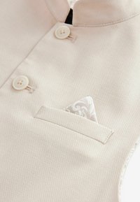 Tissu beige clair avec un motif texturé, doté d'un col montant, de deux boutons et d'un pochette en motif blanc.