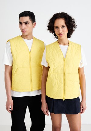 Yellow quilted vests with a zipper, featuring a textured pattern, armholes, and front pockets, worn over simple white tops.